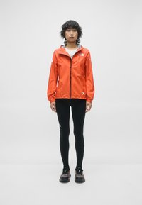 Veste légère rouge avec capuche, doublure grise contrastante, leggings noirs et chaussures de trail noires avec des accents. Figure debout, fond neutre.