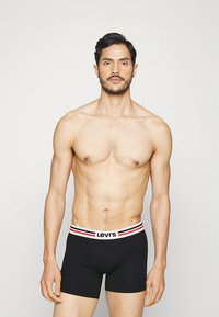 Črne boksarice z rdeče-belim elastnim pasom s črtami, na katerem je logo Levi's. Gladek material in prilegajoč dizajn.