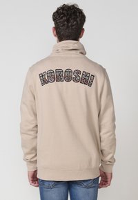 Felpa con cappuccio beige caratterizzata da una grande grafica "KOROSHI" multicolore sul retro, con una texture morbida e polsini a coste.