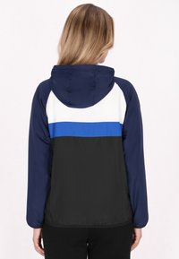 Vindjakke i navy, hvid og sort. Har hætte, elastiske manchetter og farveblokdesign med et blåt stribet mønster på ryggen.