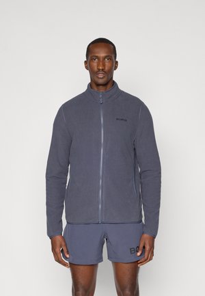 Sivá fleecová bunda s vysokým golierom, predným zipsom a jemným logom. Nosená over zodpovedajúcich šortkách, ktoré ukazujú hladkú textúru.