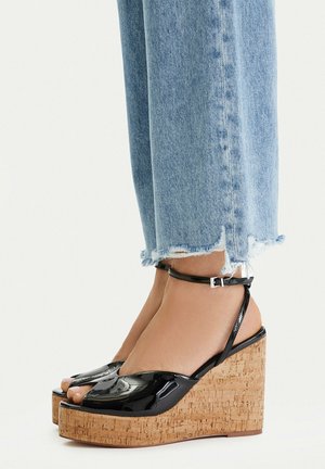 Piedi che indossano sandali con zeppa open-toe in pelle verniciata nera con cinturini alla caviglia, abbinati a jeans cropped a lavaggio chiaro con orlo sfrangiato.