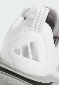 Gros plan sur le talon d'une chaussure de sport blanche avec un logo Adidas cousu et une languette avec des bandes réfléchissantes au-dessus de la semelle texturée.