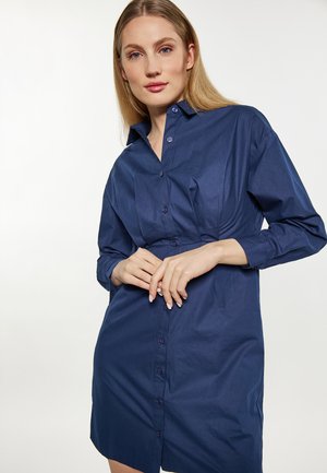 Femme aux cheveux blonds et raides portant une robe-chemise bleu marine à manches longues avec col, posant les mains jointes à la taille.