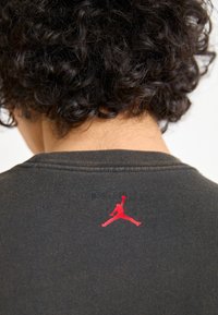 Camiseta de color gris oscuro hecha de algodón, con un pequeño logo rojo de Jumpman en la parte posterior cerca del cuello. Mangas cortas y diseño sencillo.