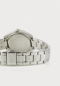 Ceas brățară din metal argintiu cu link-uri, cu închizătoare gravată "GUESS", prezentat din spate pe un fundal alb.