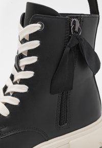 Stivaletto in pelle nera con lacci bianchi, zip laterale e un accento a nastro. Presenta una parte superiore testurizzata e una suola spessa color crema.