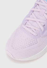 Zapato deportivo de color púrpura claro con parte superior texturizada, cordones suaves y puntera reforzada. Detalles de malla destacables y suela blanca con motas.