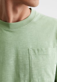 Hellgrünes Baumwoll-T-Shirt mit Rundhalsausschnitt, strukturierten Oberflächen und einer aufgenähten Brusttasche auf der linken Seite. Verfügt über ein minimalistisches Etikettendetail.