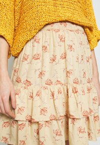 Gonna avvolgente floreale in tessuto beige chiaro con motivi di fiori rossi, caratterizzata da un orlo arricciato e una vita elastica per il massimo comfort.