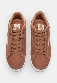 Sneaker in suede marrone con lacci piatti, suola in gomma bianca e dettagli color sabbia. Presenta un'etichetta con logo sulla linguetta e una fodera in tessuto testurizzato.