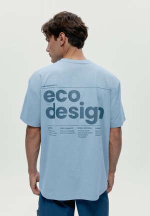 Mladý muž s krátkymi hnedými vlasmi, oblečený v voľnom svetlomodrom tričku s potlačou "eco design" a princípmi udržateľnosti na chrbte v tmavomodrej farbe.