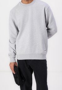 Sweat-shirt gris à manches longues, col rond et poignets côtelés. Associé à un jean noir, tenant un petit sac noir zippé.