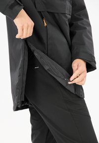 Svart ytterjacka med dragkedja framtill, med ett tygpanel och en ficka inuti. Jackan är lager-på-lager över svarta byxor.