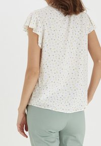 Leichte weiße Bluse mit kleinen gelben und pinkfarbenen Blumenmustern. Verfügt über kurze, gerüschte Ärmel und einen abgerundeten Saum.