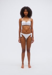 Σετ λευκό μαγιό μπικίνι με τοπ bandeau με το λογότυπο 'EA7' και κάτω μέρος με κορδόνια στο πλάι. Λείο υλικό και μινιμαλιστικός σχεδιασμός. Το μοντέλο στέκεται ξυπόλητο.