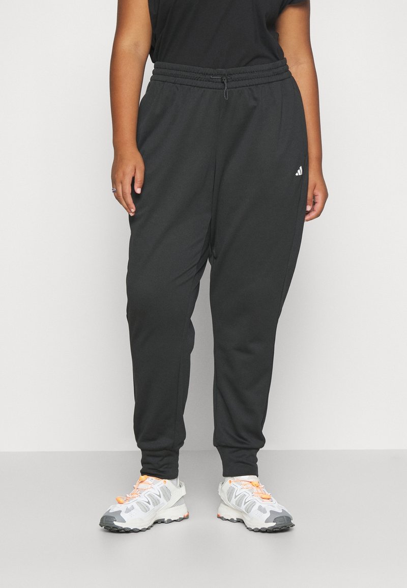 Personne portant un pantalon de survêtement noir Adidas et des baskets blanches avec des lacets orange, se tenant contre un fond clair.