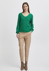 Grüner Strickpullover mit V-Ausschnitt und strukturiertem Muster, kombiniert mit beigen, schmal zulaufenden Hosen und spitzen Stiefeletten.