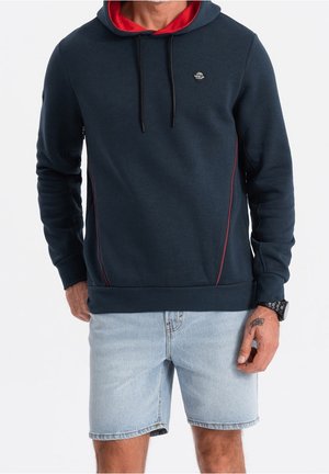 Mann trägt marineblauen Hoodie mit roter Innenfütterung und hellblauen Denim-Shorts, zeigt Tätowierung an der rechten Hand und schwarze Armbanduhr.