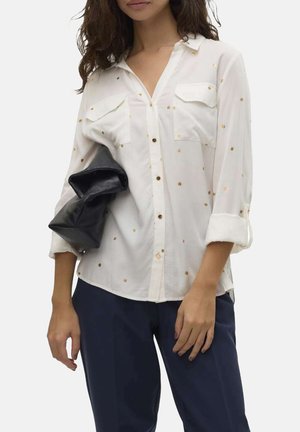 Chemise blanche à boutons avec un motif de points dorés, dotée de deux poches avant et de manches retroussées, associée à un sac noir et un pantalon sombre.