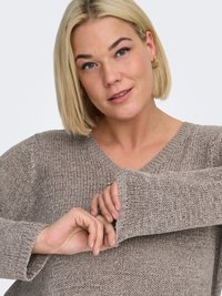 Light brown knit sweater with a V-neck and long sleeves, featuring ribbed cuffs and a textured finish. Hands partially visible near sleeves.