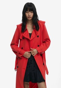 Red trench coat with a double-breasted design, large black buttons, wide lapels, and belted cuffs, layered over a black dress.