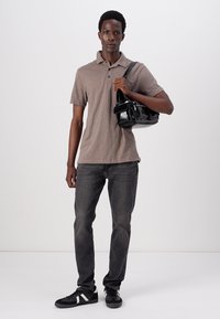 Polo à manches courtes marron, jean foncé et baskets noires. Tenant un sac bandoulière noir brillant avec plusieurs compartiments.