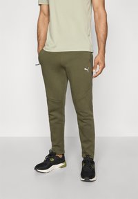 Olivgrüne Jogginghosen mit einer glatten Textur, ausgestattet mit Seitentaschen und einem weißen Puma-Logo an der linken Hüfte, getragen mit schwarzen Sportschuhen.