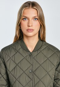 Mladá žena s modrými očami a blond vlasmi má na sebe zlaté kruhové náušnice a prešívanú olivovo zelenú bundu s cvokmi na jednoduchom pozadí.