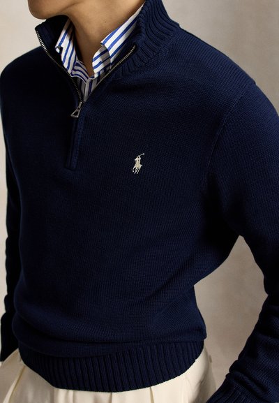 Homme portant un pull zippé bleu marine par-dessus une chemise rayée bleue et blanche à col, avec un pantalon beige visible en dessous.