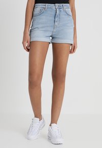Shorts de mezclilla azul claro con dobladillo vuelto, diseño de cinco bolsillos y cintura ajustada. Se usan con zapatillas deportivas blancas.