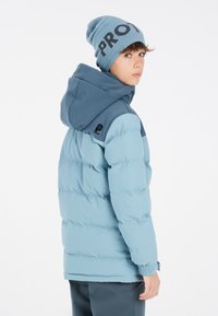 Puffige blaue Jacke mit einer dunkler blauen Kapuze, elastischen Bündchen und einem Logodetail auf der Schulter. Dazu gehört eine passende hellblaue Mütze.