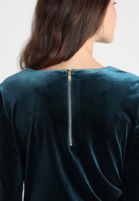 Teal sammetstopp med båturringning, lång ärm och en synlig gulddragkedja på baksidan för stängning. Slät textur med en subtil glans.