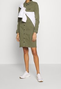 Vestido camisero de color verde oliva con botones en la parte delantera, bolsillos en el pecho, mangas largas y un sudadera blanca atada a la cintura; combinado con zapatillas blancas.