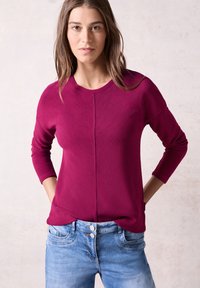 Magenta gerippter Pullover mit langen Ärmeln, rundem Halsausschnitt und diagonalen Texturdetails, kombiniert mit hellblauen Jeans.