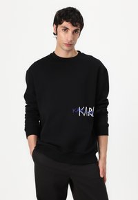 Felpa nera in cotone con colletto a girocollo, maniche lunghe e un logo ricamato in blu e bianco sul lato sinistro.