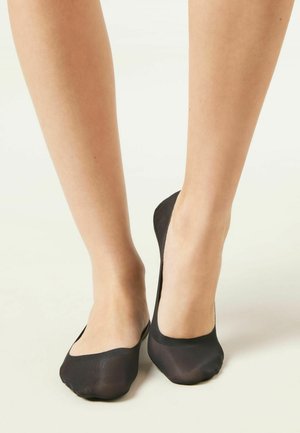 Nohy v černých průhledných ponožkách typu "no-show" na světlém pozadí, zobrazující kůži kolem kotníku a spodní části nohy.