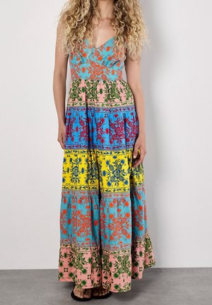 Femme portant une robe maxi sans manches à motifs floraux multicolores avec des motifs superposés en bleu, jaune, rose et vert, debout sur un sol gris.