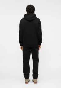 Μαύρο φούτερ με κουκούλα και τσέπη καγκουρό, υφασμένο υλικό με υφή και ριμπ μανσέτες. Συνδυάζεται με σκούρο παντελόνι και μπεζ μποτάκια, πανοραμική όψη από πίσω.