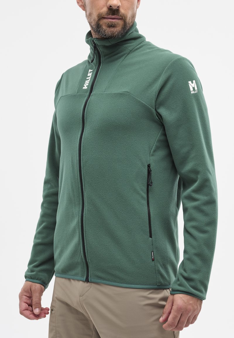 Green fleece jacket with high collar, zippered front, side pockets, textured fabric, and contrasting black zipper detail. Logo on sleeve.