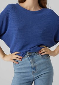 Pull tricoté bleu avec un large col et un motif texturé, porté avec un jean en denim bleu clair taille haute.