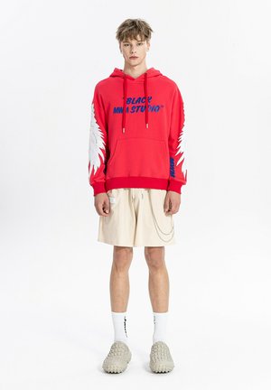 Joven con sudadera roja con diseños de alas blancas en las mangas, pantalones cortos beige con cadena, calcetines blancos y zapatos beige texturizados, de pie recto.