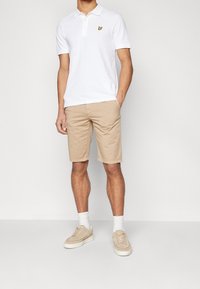 Mann trägt ein weißes kurzärmliges Poloshirt mit gelbem Vogel-Logo, beige Shorts, weiße Socken und beigen Sneakers und steht mit einer Hand in der Tasche.