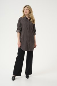 Chemise grise à boutons avec un tissu texturé, une poche poitrine et des manches retroussées, associée à un pantalon noir à jambes larges et des chaussures noires à bouts pointus.