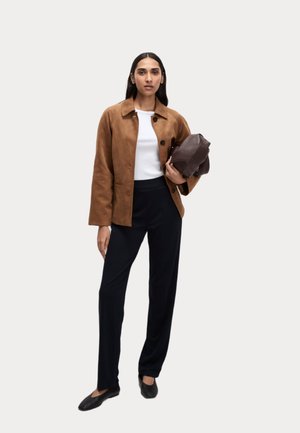 Veste en daim marron avec des boutons noirs, haut blanc et pantalon sombre. Le mannequin tient un sac marron foncé et porte des ballerines noires. Fond neutre.
