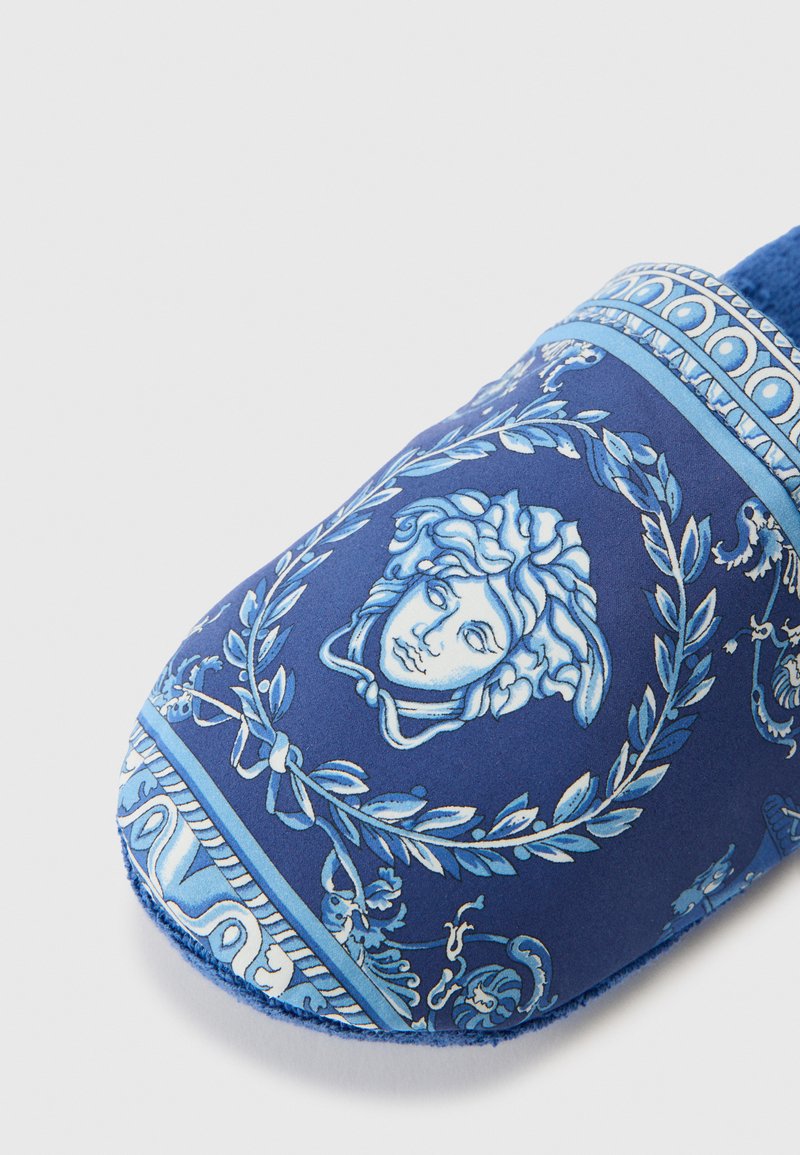 Blaue Hausschuhe mit einem detaillierten Muster, das ein Gesichtsmotiv, blattförmige Akzente und hellere blaue Highlights zeigt. Weicher Stoff mit abgerundetem Zehendesign.