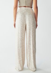Pantaloni a palazzo in beige chiaro con texture luccicante. Presentano una vita alta e una vestibilità ampia, creando una silhouette fluida.