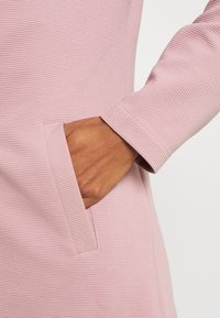 Jasnoróżowa fakturowana tkanina z pionowym prążkowaniem, z boczną kieszenią oraz dopasowanym rękawem z czystym wykończeniem mankietu.