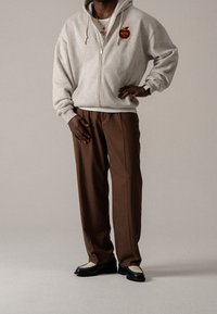 Szara bluza z kapturem zapinana na zamek z grafiką czerwonego jabłka, brązowe spodnie i czarne buty z kremowymi akcentami. Gładka tkanina i luźny krój.