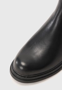 Primer plano de la punta de una bota de cuero negro con textura suave y suela cosida sobre un fondo blanco.